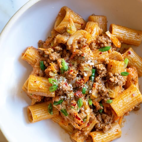 Steaming bowl of Chaos Cooking Kimchi Ragu Pasta, with rich red sauce, garnished with green scallions.